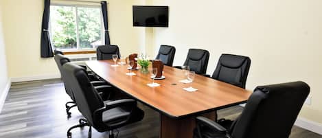 A conference room with a long table, six chairs, a television on the wall, and a window with curtains.