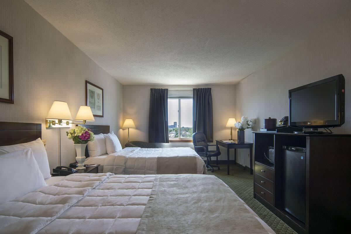 A hotel room with two beds, a desk, a television, and a view of the city.