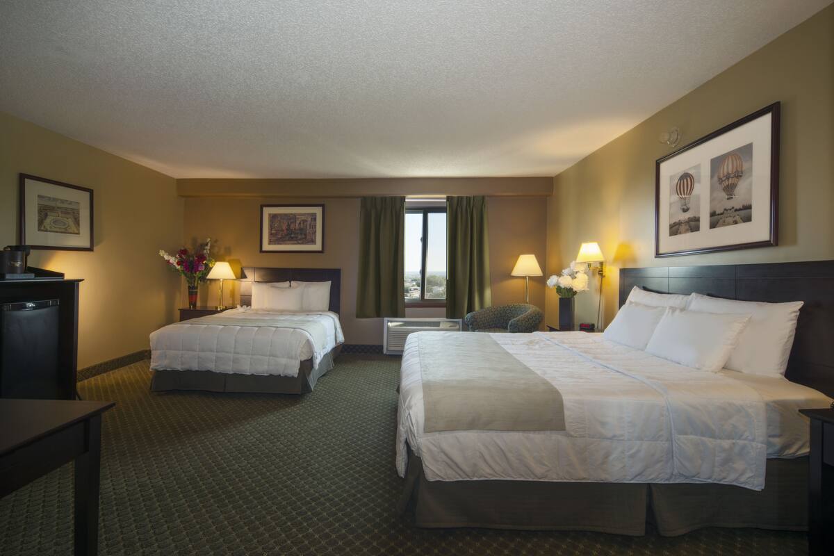 A hotel room with two beds, a desk, a chair, a window with curtains, and wall-mounted lamps.