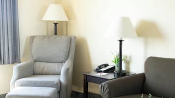 A hotel room with a grey armchair, a small table with a lamp, a telephone, and a vase with flowers.