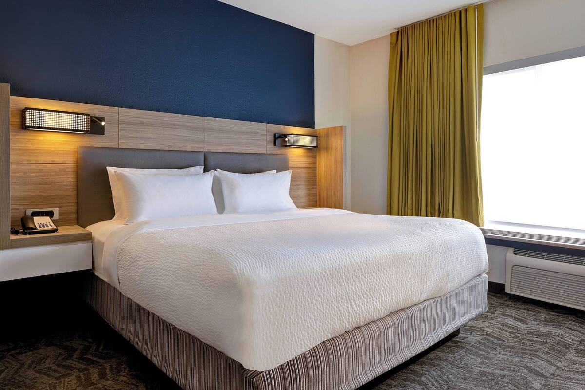 A hotel room with a large bed, a nightstand, a wall-mounted light, and a window with curtains.