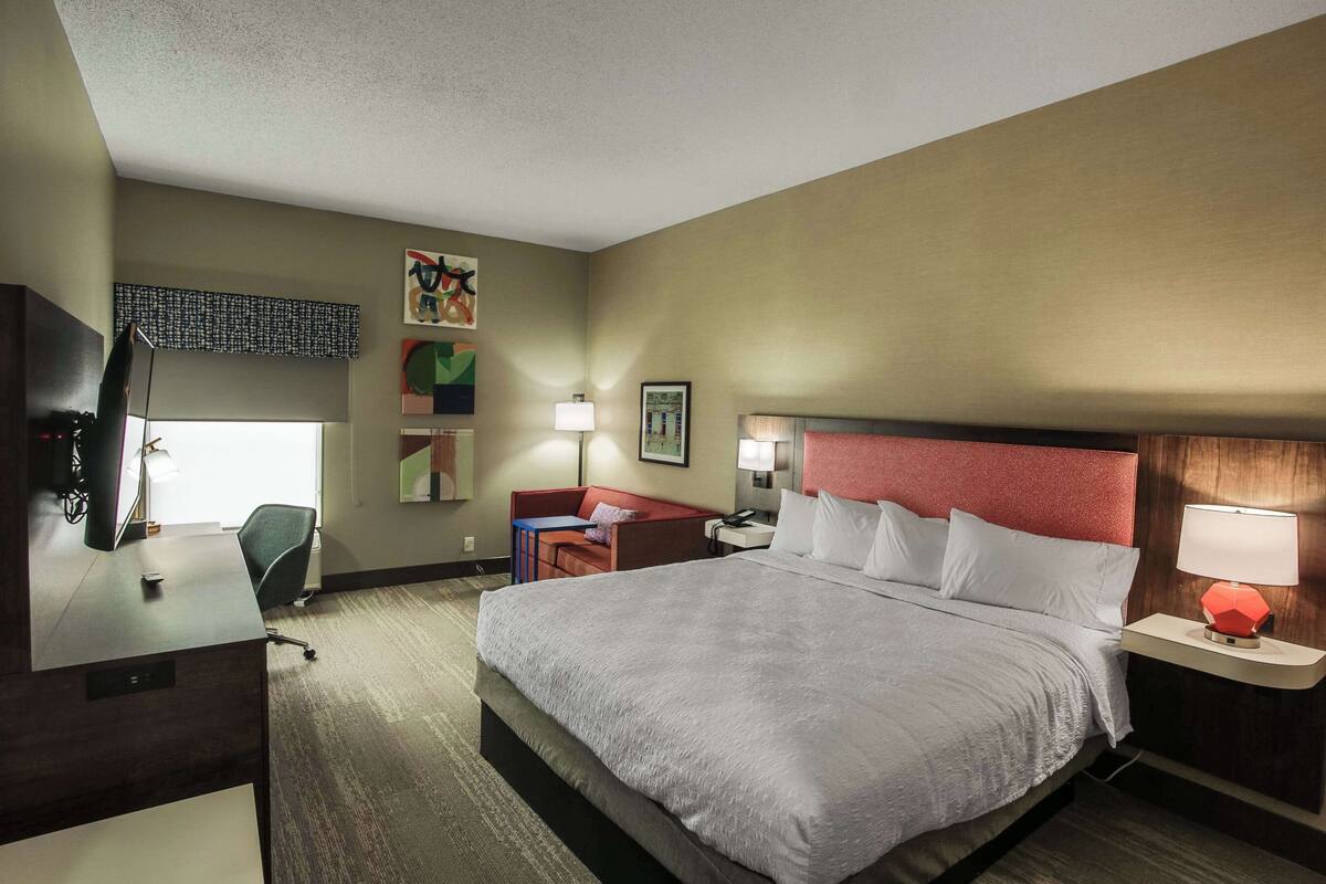 A hotel room with a large bed, a desk with a computer, a chair, a desk lamp, a nightstand with a lamp, and a window with blinds.