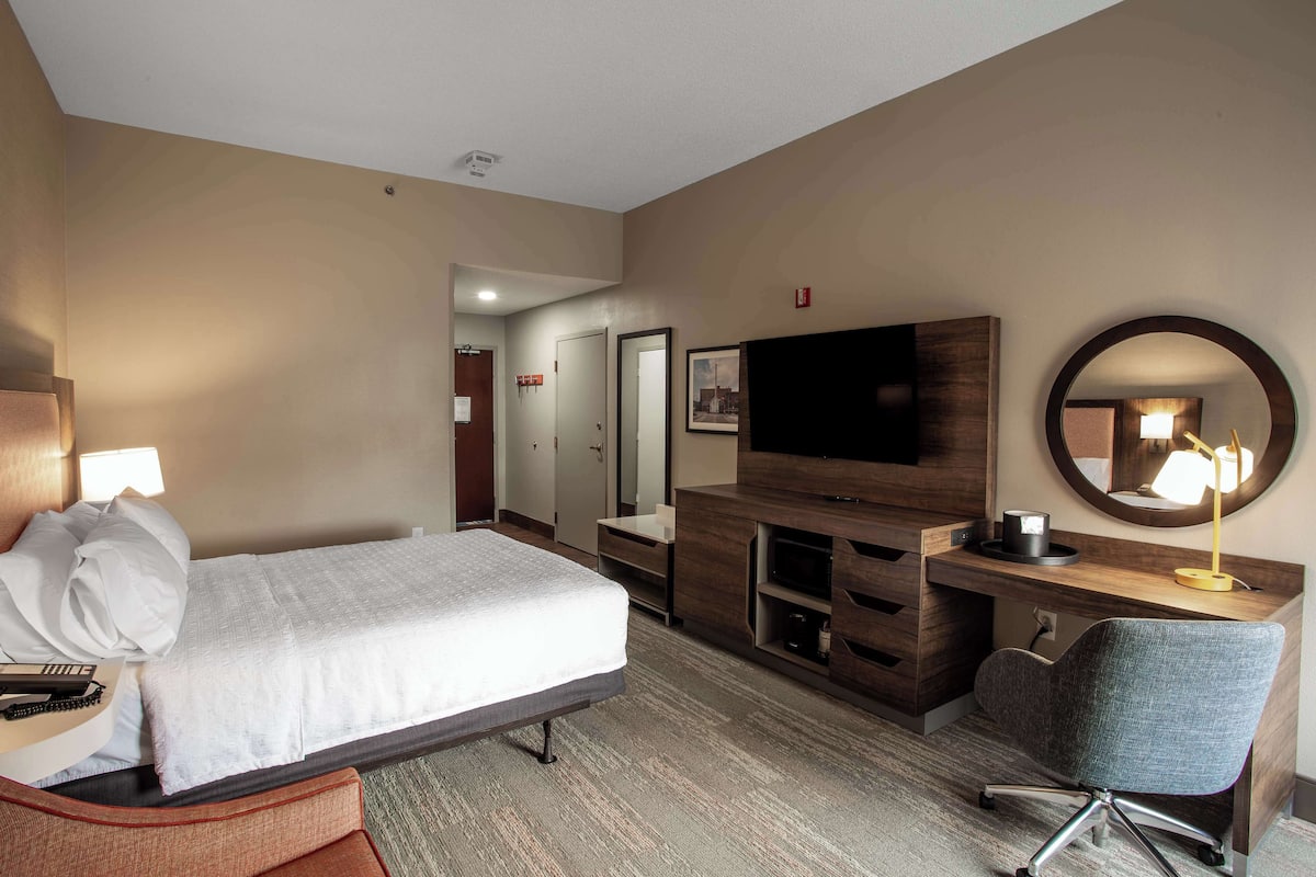 A hotel room with a bed, a desk with a chair, a television, a mirror, and a lamp.