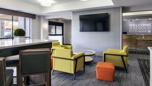 A modern lobby with a bar area, seating with yellow patterned chairs, a flat-screen TV mounted on the wall, and a welcome sign. - Hampton Inn Springfield (Springfield)