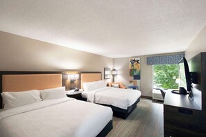A hotel room with two beds, a desk with a computer, a TV, and a window with a view of trees.