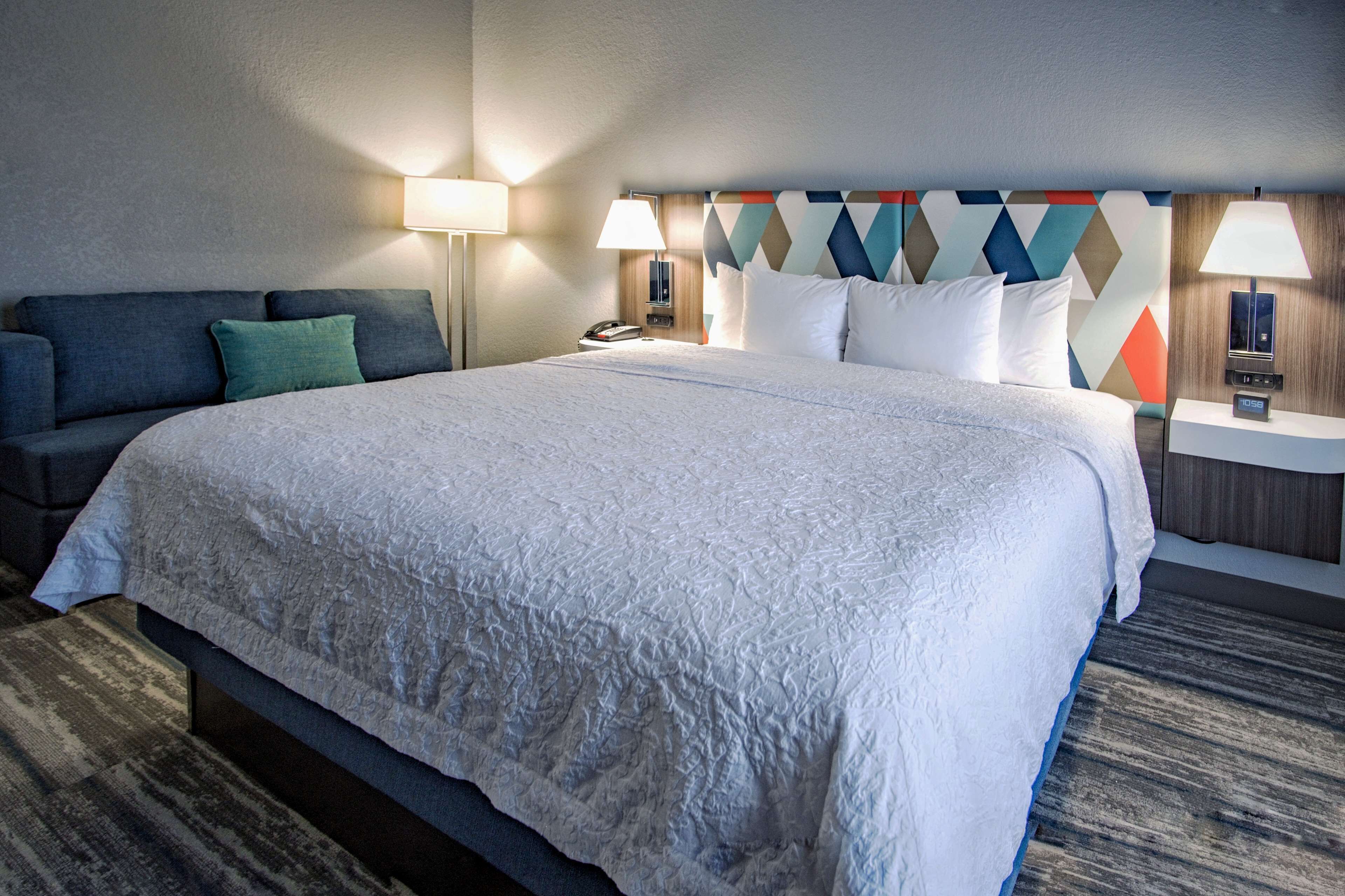 A hotel room with a large bed, a sofa, two bedside tables with lamps, and a geometric-patterned headboard.