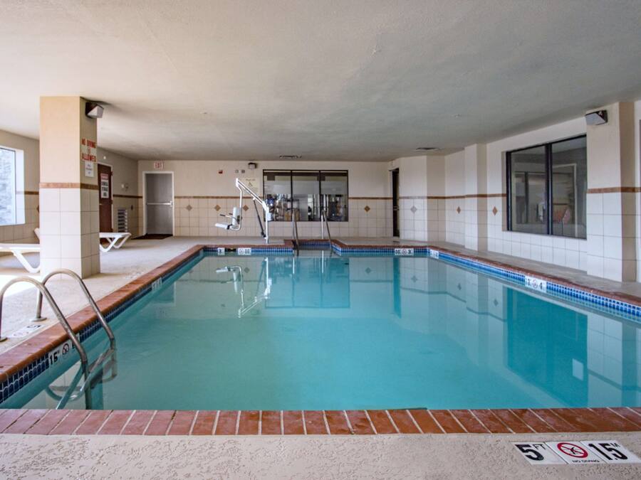 Una piscina con agua azul cristalina, paredes con azulejos y un borde de ladrillo. Se observan sillas de descanso y una escalera que da acceso a la piscina.