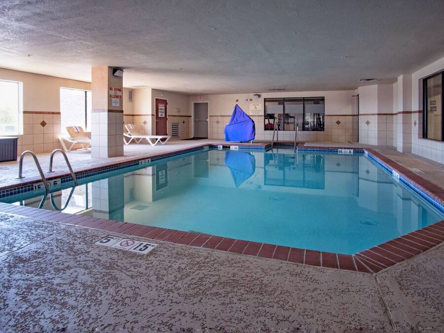 An indoor swimming pool with clear water, tiled walls, and a tiled floor. There are lounge chairs and a covered area.