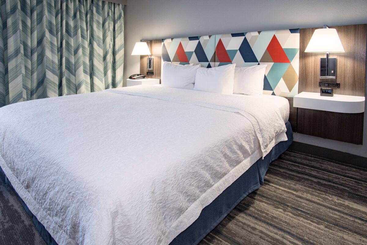 A hotel room with a large bed, two bedside tables with lamps, a modern geometric-patterned headboard, and a patterned curtain.