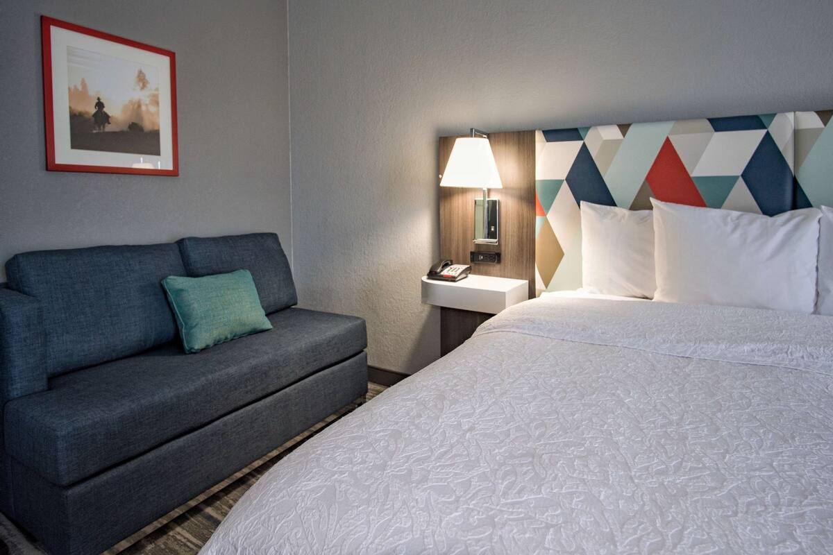 A hotel room with a bed, a sofa, a nightstand with a lamp, and a framed picture on the wall.