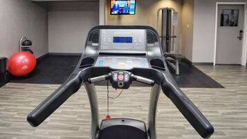 A hotel fitness center with a treadmill, a red exercise ball, and a television screen displaying a news program.