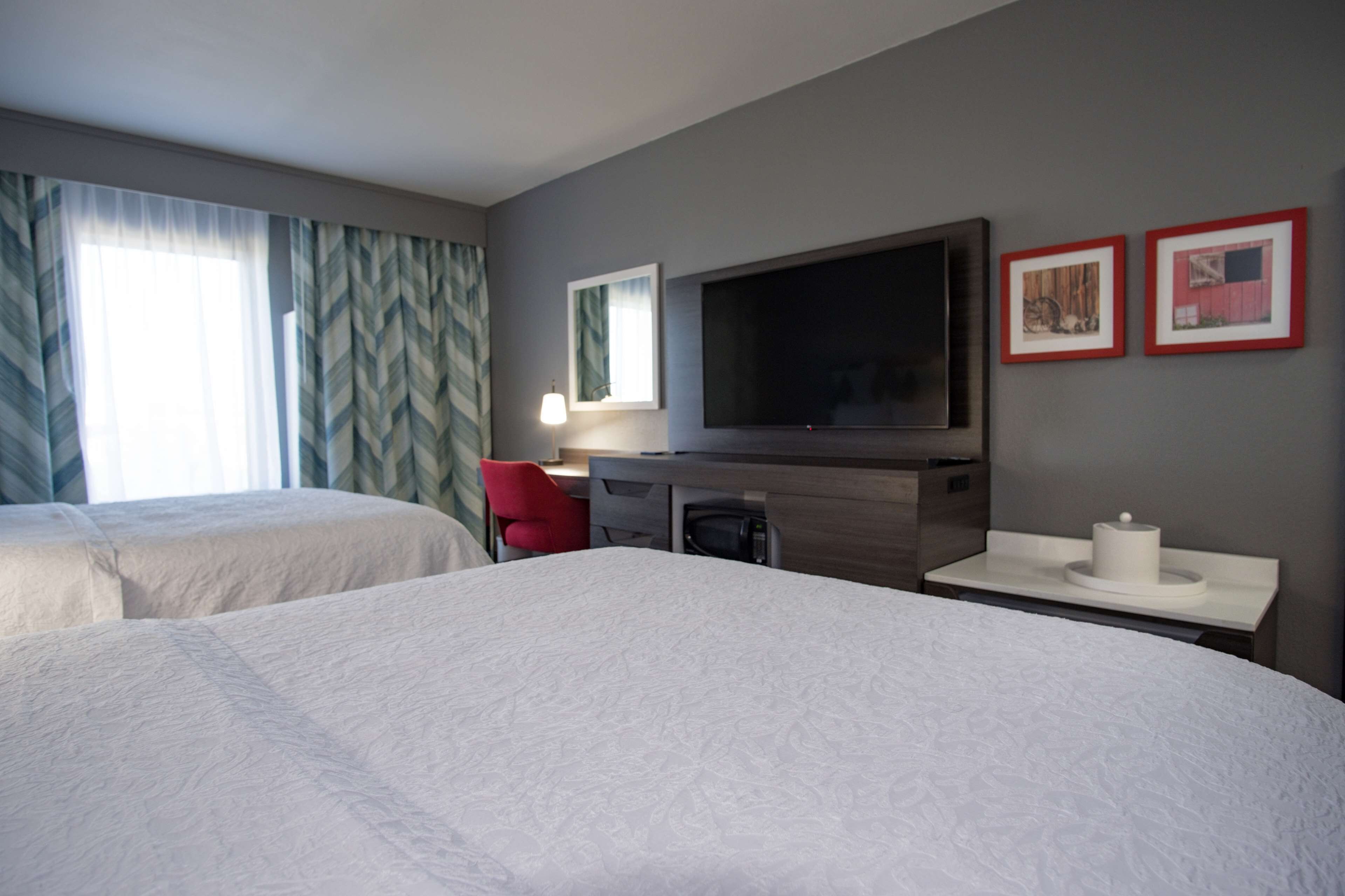 Quarto de hotel com duas camas, uma escrivaninha com televisão, uma cadeira vermelha e uma janela com cortinas.