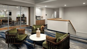 A modern hotel lobby with a seating area featuring two sofas and a coffee table, a kitchen area, and a staircase leading to an upper floor.