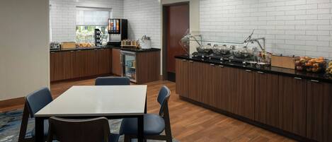 Habitación de hotel moderna con zona de comedor que incluye una mesa y sillas, kitchenette con refrigerador y máquina de café, y barra de desayuno con fruta y aderezos.