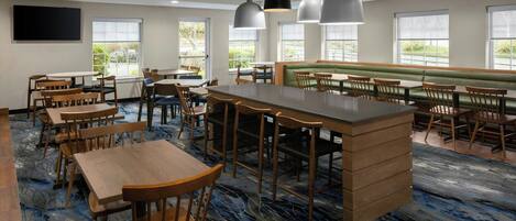 A spacious dining area with wooden tables and chairs, a central bar, and large windows providing natural light.