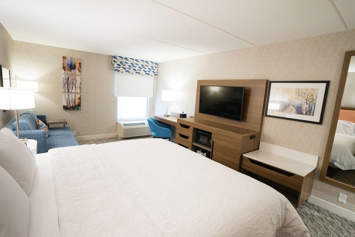 A hotel room with a large bed, a desk with a flat-screen TV, a chair, and a window with blinds.
