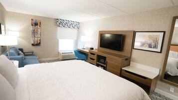 Een hotelkamer met een groot bed, een bureau met een flatscreen televisie, een stoel en een raam met jaloezieën.