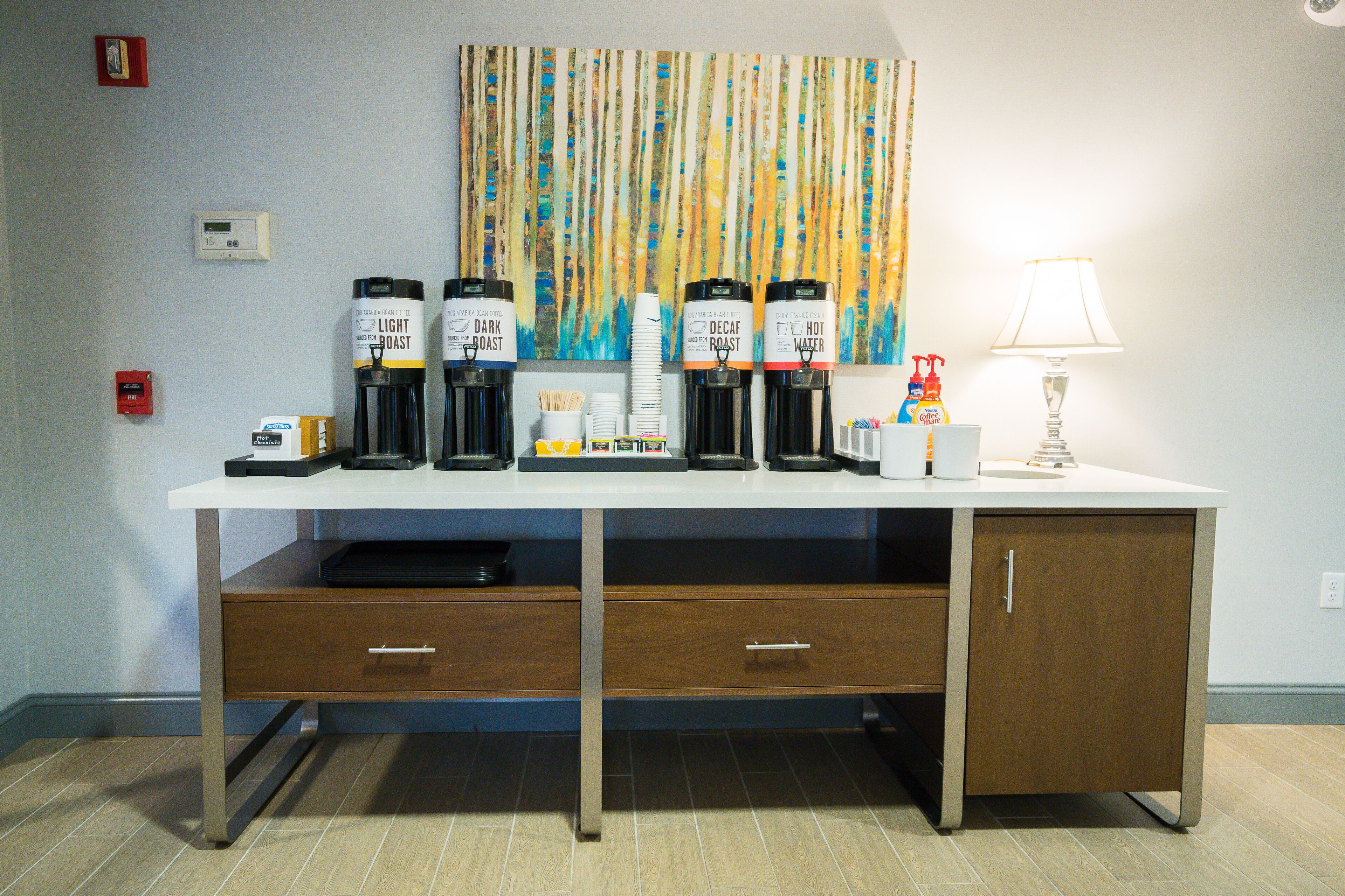 Een hotelkamer met een bureau waarop koffieautomaten staan, gelabeld 'Light Roast', 'Dark Roast', 'Decaf Roast' en 'Warm Water'. Er zijn twee lades, een lamp en een rookmelder aan de muur.