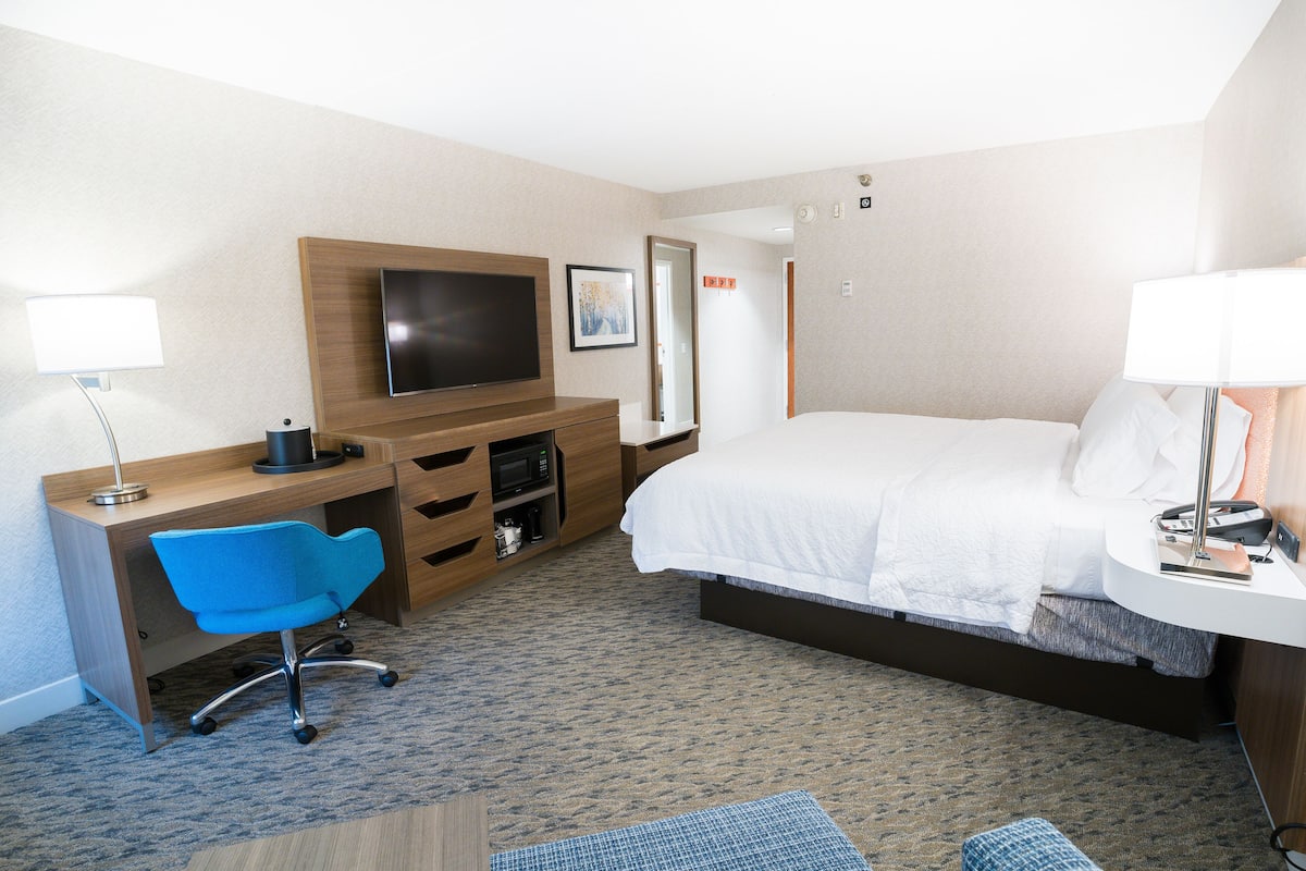 A hotel room with a bed, a desk with a chair, a TV, and a bedside table with a lamp.