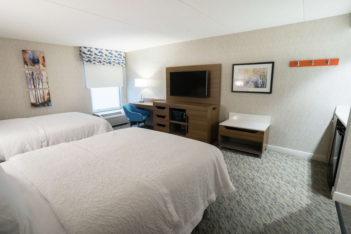 A hotel room with two beds, a desk with a chair, a flat-screen TV mounted on the wall, a small table, and a window with a patterned curtain.