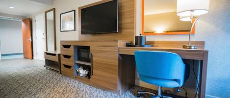 Een hotelkamer met een houten bureau, een blauwe bureaustoel, een flatscreen televisie, een spiegel en een lamp.
