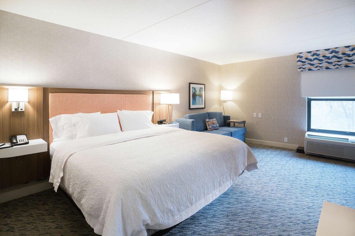 A hotel room with a large bed, bedside lamps, a small table, a chair, and a window with an air conditioner.