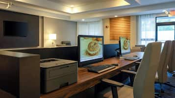 Escritório de hotel com dois monitores de computador, uma impressora e uma mesa com cadeiras.