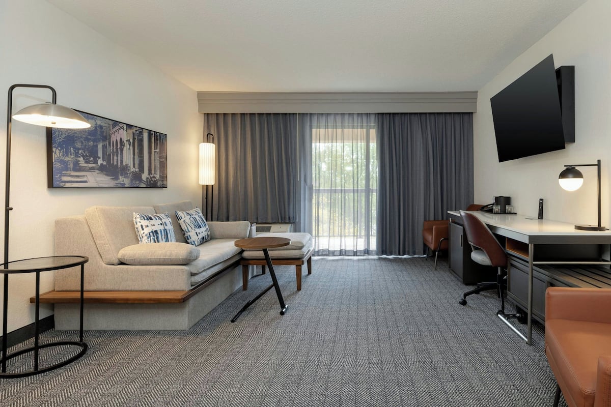 Ein Hotelzimmer mit einer Couch, einem kleinen runden Tisch, einem Schreibtisch mit Stuhl, einem an der Wand montierten Fernseher und einem großen Fenster mit Vorhängen.