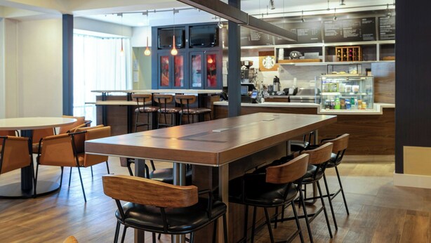 Un restaurant moderne avec une longue table en bois et des chaises noires, un espace cuisine équipé d’un réfrigérateur, et un bar avec des tabourets.