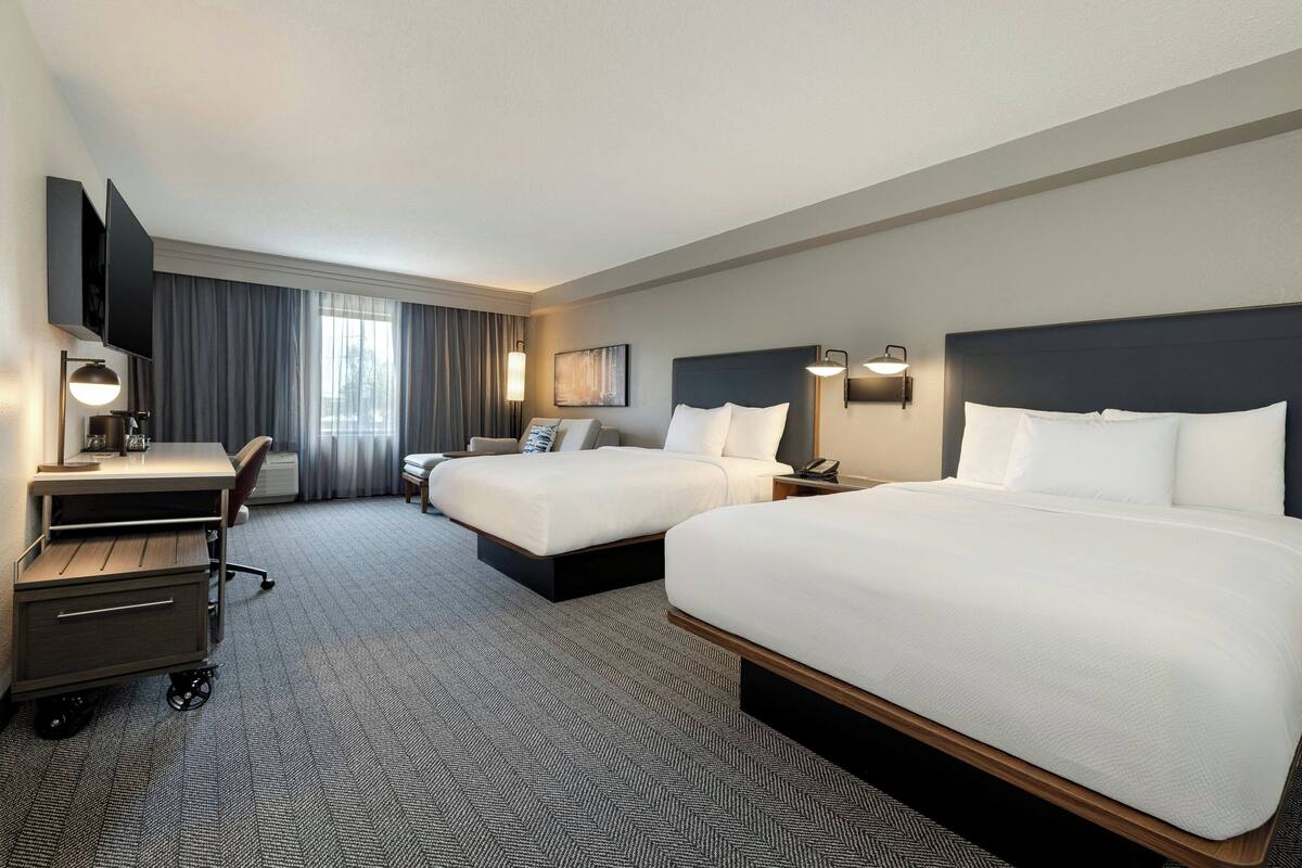 Ein Hotelzimmer mit zwei Betten, einem Schreibtisch, einem Sessel, einem Fernseher und einem großen Fenster mit Vorhängen.