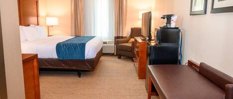 A hotel room with a bed, a desk, a chair, a bench, a TV, and a coffee machine.