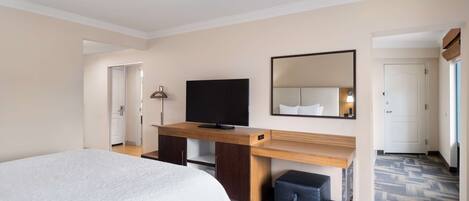 A hotel room with a bed, a desk with a TV, a mirror, and a door.