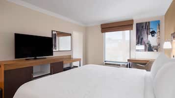 A hotel room with a large bed, a flat-screen TV on a wooden desk, a window with a view of buildings, and a painting on the wall.