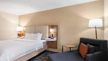 A hotel room with a bed, a bedside table with a lamp, a small table, and a sofa with a pillow.