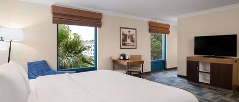 Quarto de hotel com uma cama grande, uma mesa, uma cadeira, uma televisão e janelas com persianas.