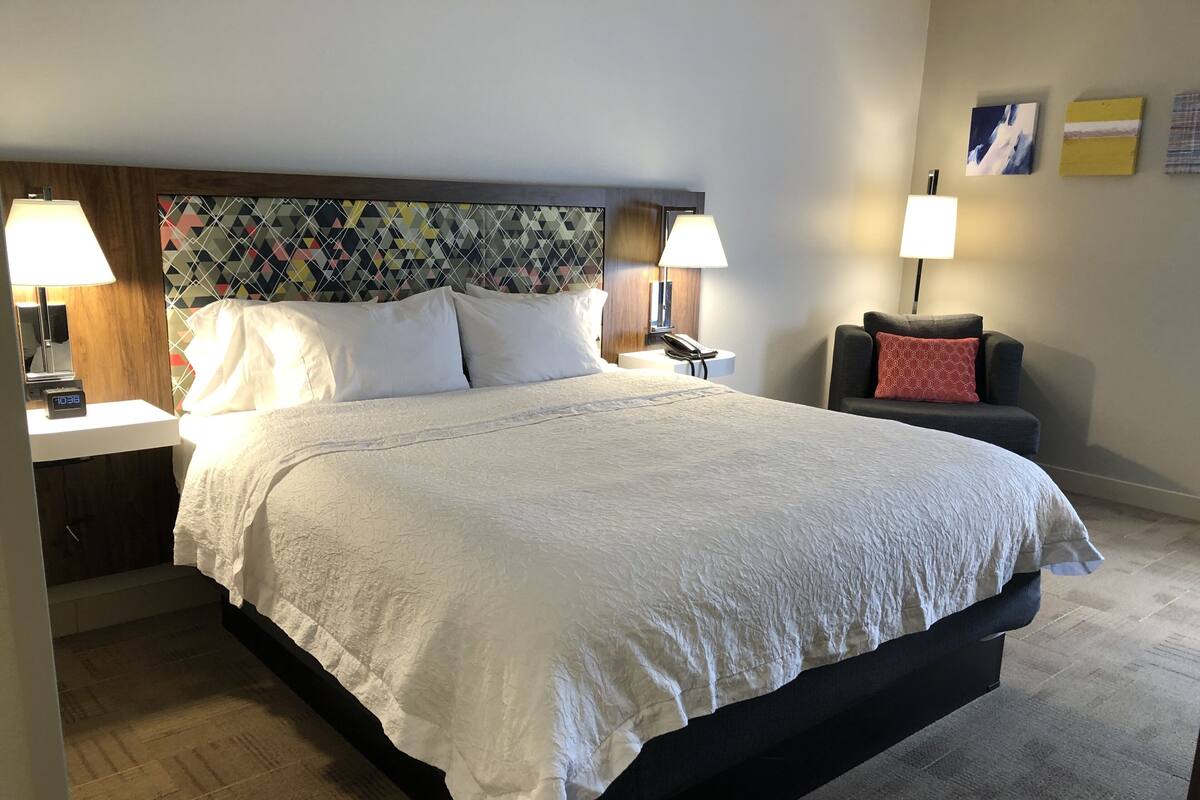 A neatly made bed with white linens, a wooden headboard with a geometric pattern, two bedside lamps, a nightstand with a phone, and a small table with a lamp.