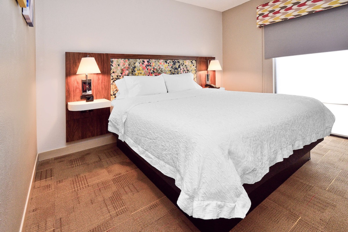 A hotel room with a large bed, two bedside tables with lamps, a patterned headboard, and a window with a view.