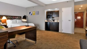 Habitación de hotel con cama, escritorio, silla, lámpara, kitchenette y puerta.