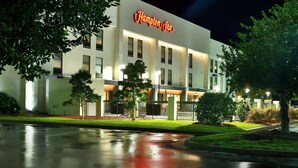 Un hotel Hampton Inn de noche, con una fachada iluminada y un estacionamiento al frente.