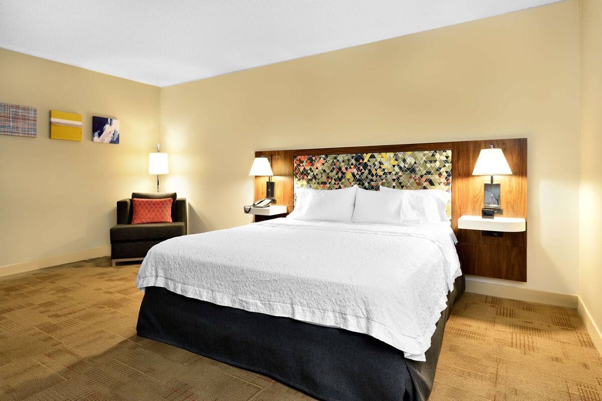Una habitación de hotel con una cama grande, dos mesitas de noche con lámparas, un sofá con un cojín rojo y una pared con tres cuadros enmarcados.