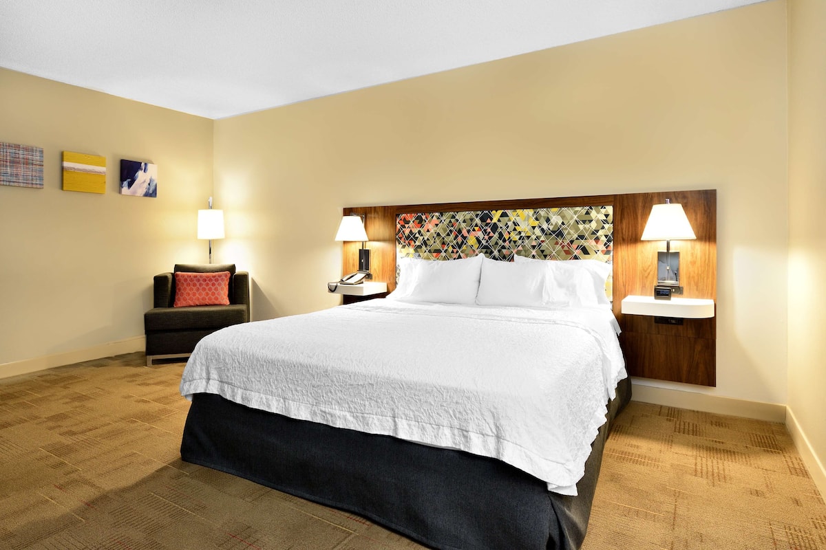 A hotel room with a large bed, two bedside lamps, a nightstand, a chair with a red cushion, and a wall with three framed pictures.