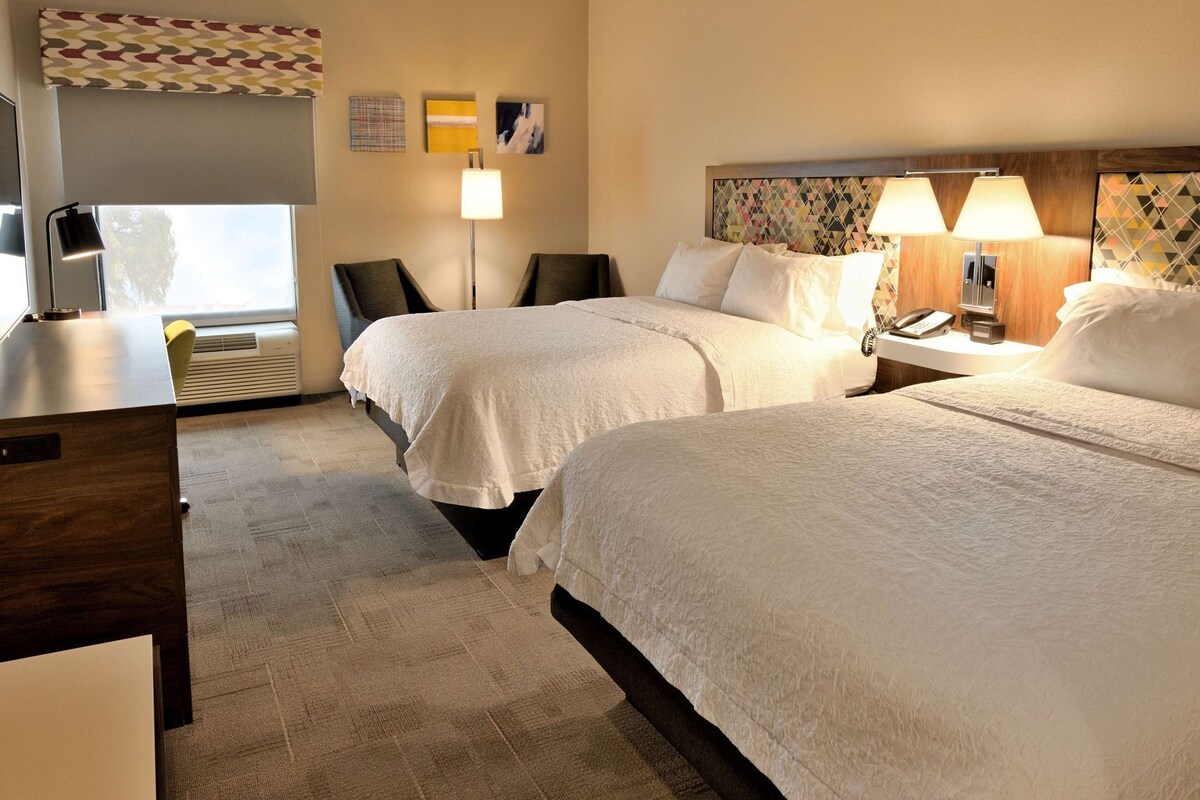 A hotel room with two beds, a desk, a chair, a lamp, and a wall with artwork.