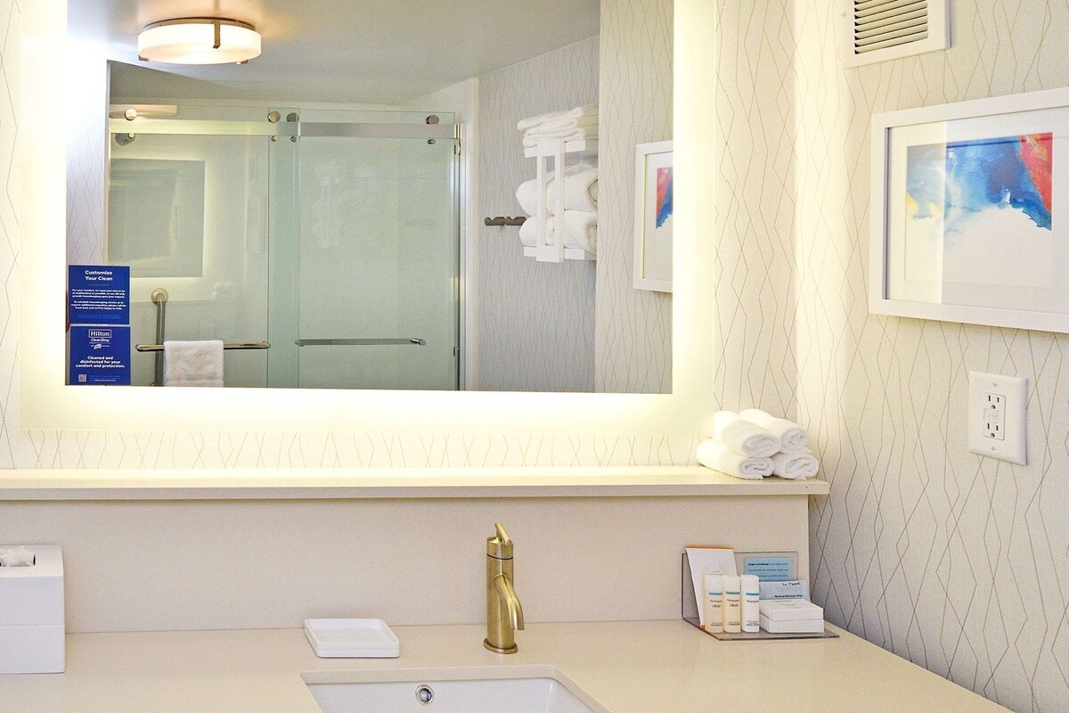 A hotel bathroom with a large mirror, a sink, a towel rack, and a framed artwork on the wall.