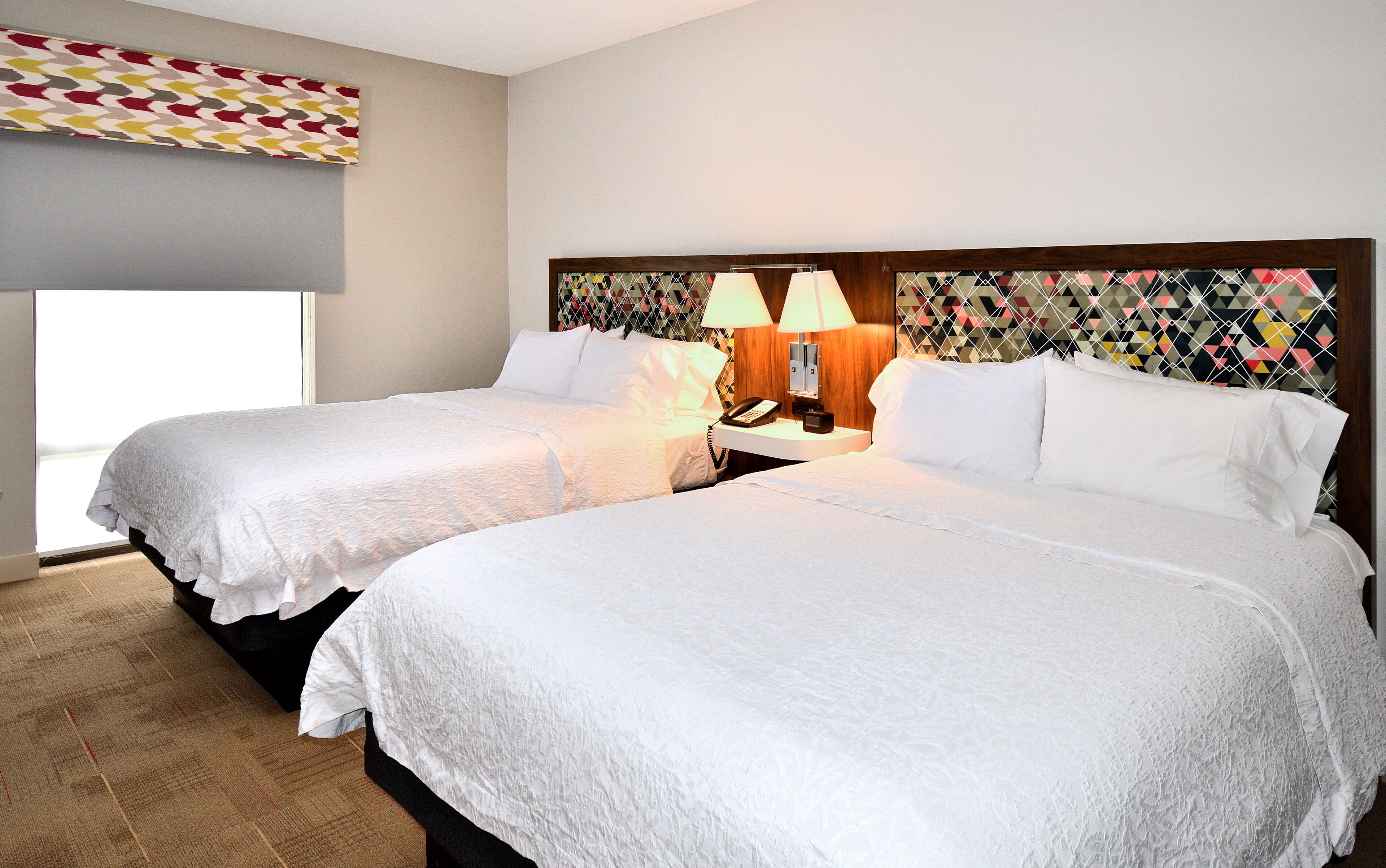 A hotel room with two beds, a nightstand with a lamp, and a patterned headboard.