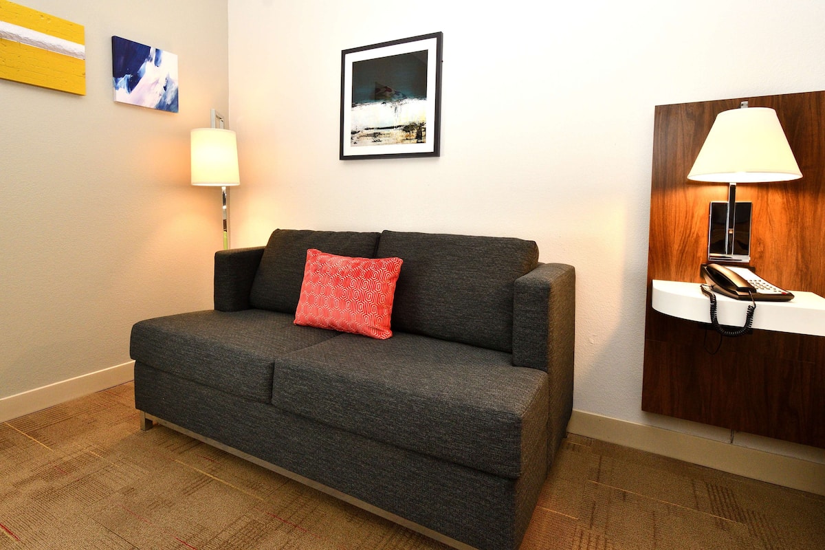 A grey sofa with a red pillow, a standing lamp, and a framed picture on the wall.