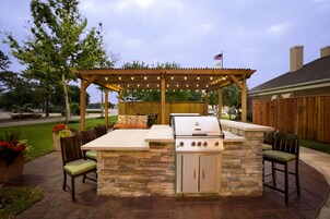 Un patio al aire libre con una estación de barbacoa de piedra, asientos y una pérgola con luces.