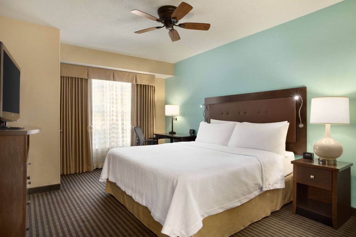 Ein Hotelzimmer mit einem großen Bett, einem Deckenventilator, einem Nachttisch mit Lampe, einem Schreibtisch mit Stuhl und einem Fenster mit Vorhängen.