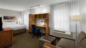 Habitación de hotel con cama, escritorio, silla, sofá, televisor, lámpara y ventana con cortinas.