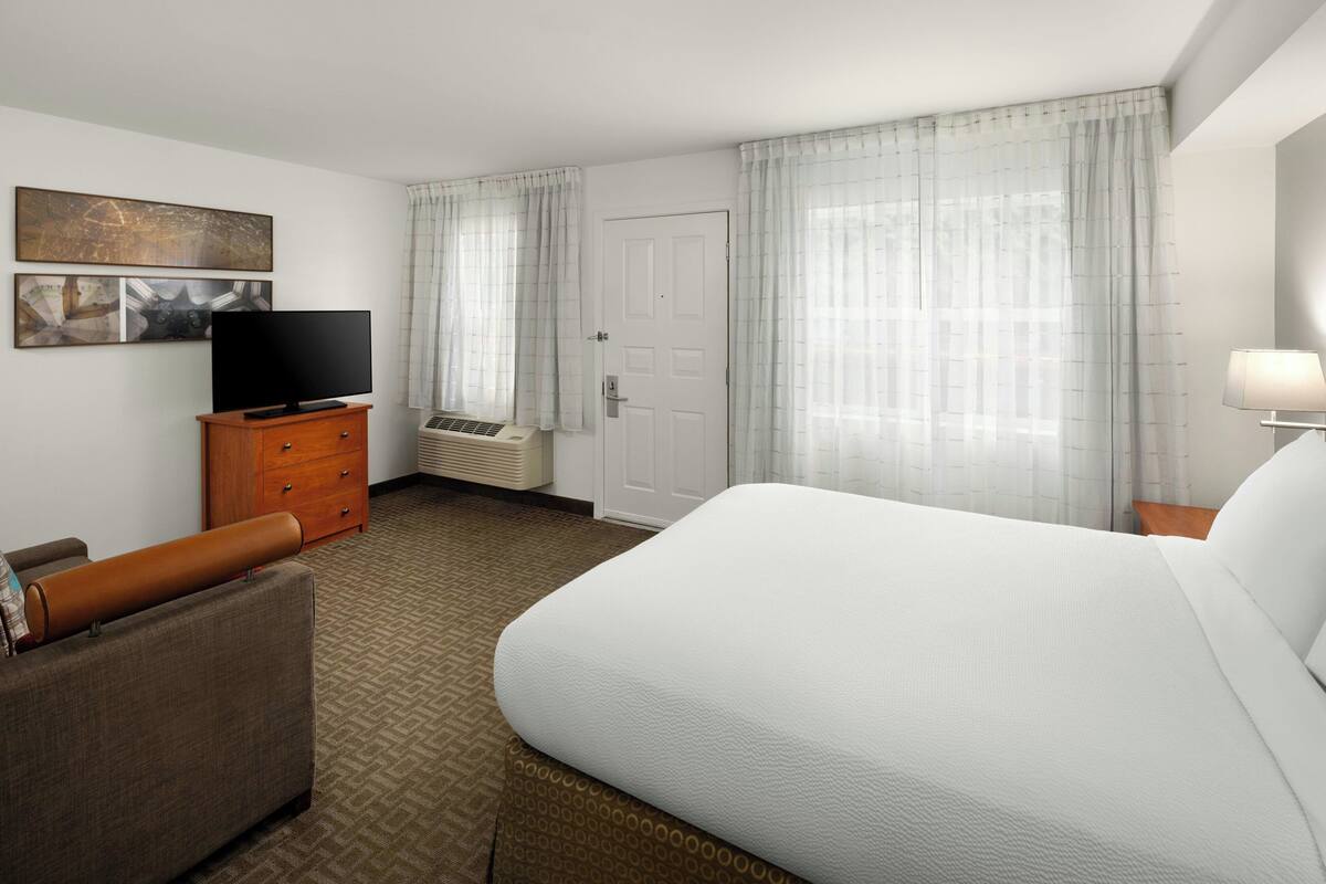 A hotel room with a bed, a TV on a wooden dresser, a desk lamp, and a window with curtains.