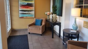 Un hall d’entrée avec un fauteuil beige, une table noire ornée d’un vase de fleurs, un tableau et un panneau indiquant la sortie.